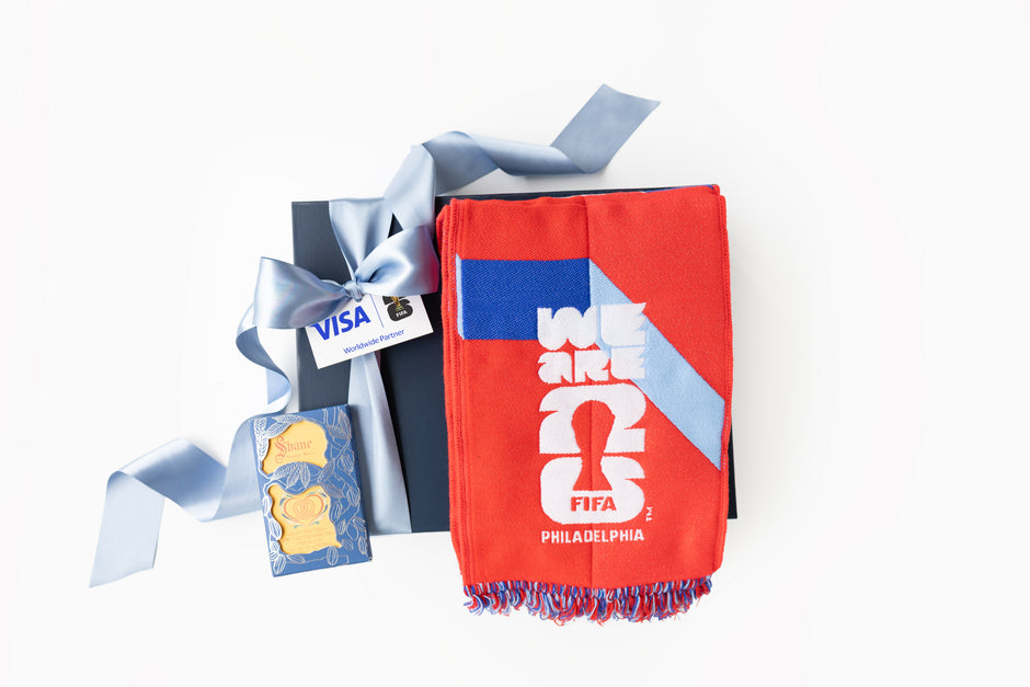 A red and blue Philadelphia soccer-themed scarf with 'WE ARE 2026 FIFA PHILADELPHIA' printed on it, presented with a dark blue gift box tied with a light blue ribbon and a small card on a white background.
