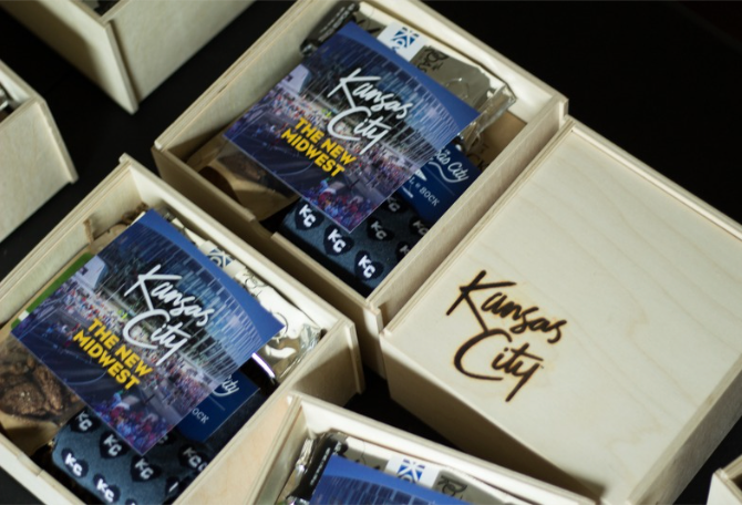 Open wooden gift boxes containing printed Kansas City gift inserts and locally sourced gifts, with one box lid engraved or printed 'Kansas City'.