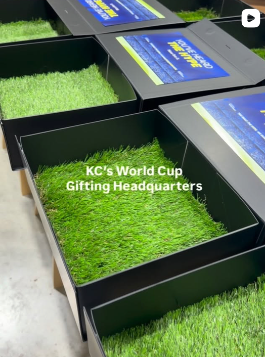 Several black rectangular boxes filled with bright green artificial grass are arranged on a concrete floor. Blue and yellow labels on the boxes read 'KC's World Cup Gifting Headquarters'.