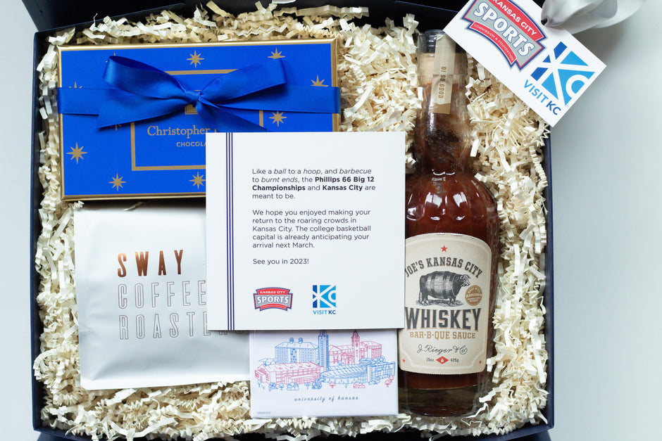 A curated gift box filled with shredded paper packaging, containing a bottle of whiskey bbq sauce, a bag of coffee, a blue chocolate box from Christopher Elbow, a printed card, and a hanging Big 12 and Visit Kansas City tag.