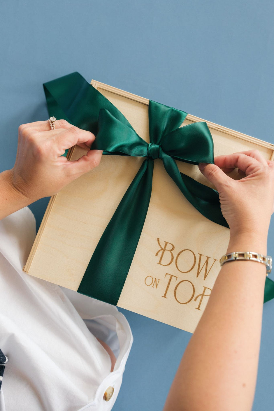 A person's hands are shown tying a large dark green satin bow onto a light wood gift box with gold lettering that reads 'BOW ON TOP'. The person is wearing a white collared shirt, and the background is a solid slate blue.