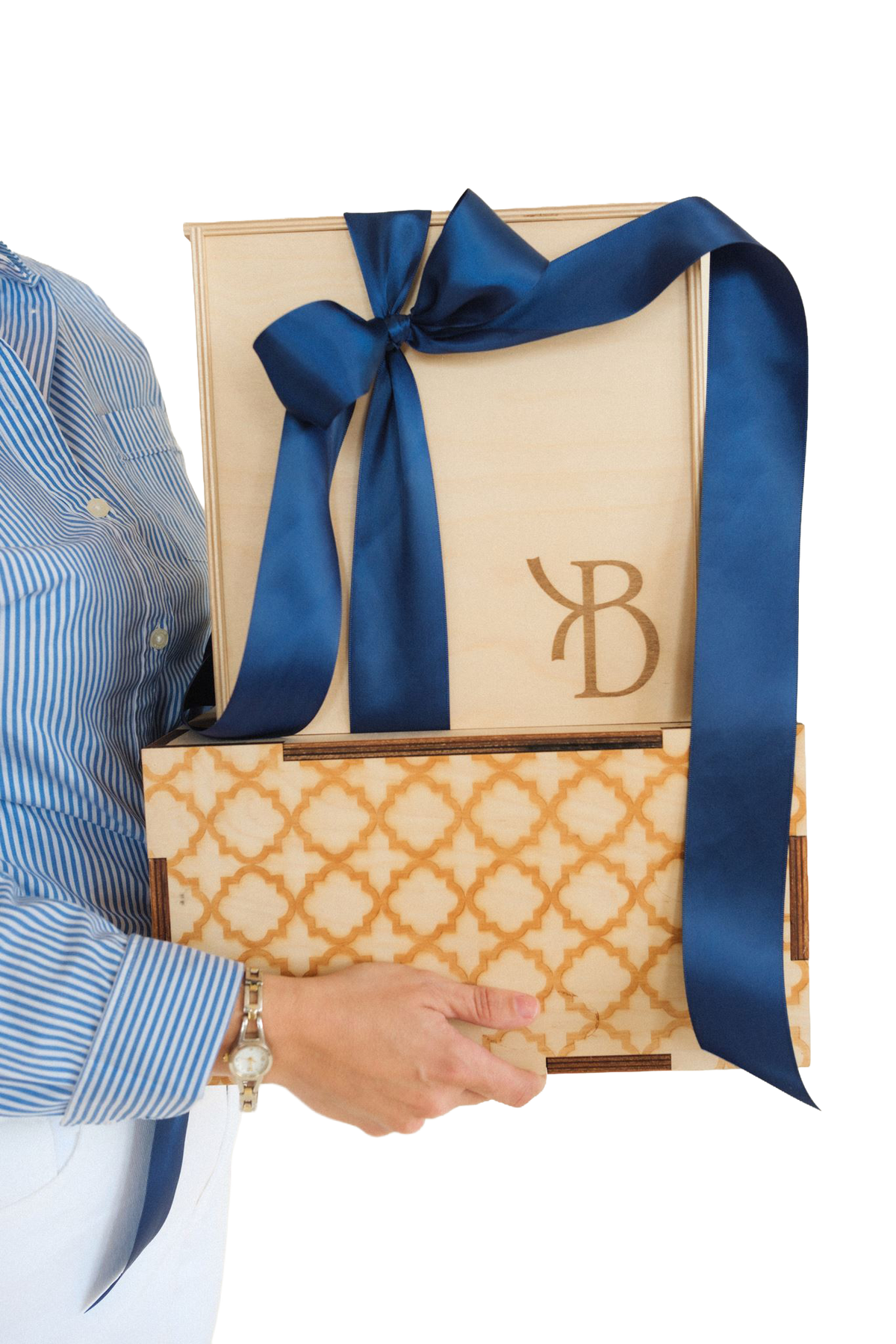 A person in a blue-and-white striped shirt holds a wooden presentation box with a large navy ribbon bow and a patterned lower box. The top box has an engraved monogram logo.